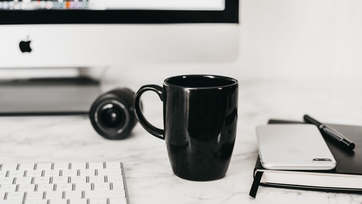 A mug of coffee on a desk beside a laptop and notepad